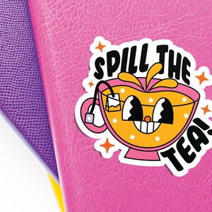 May include: A die-cut sticker featuring a cartoon teacup with a pink base and orange liquid, displaying a smiling face with large eyes. The text 'SPILL THE TEA!' is printed in bold black letters above and below the cup, accented by orange stars.
