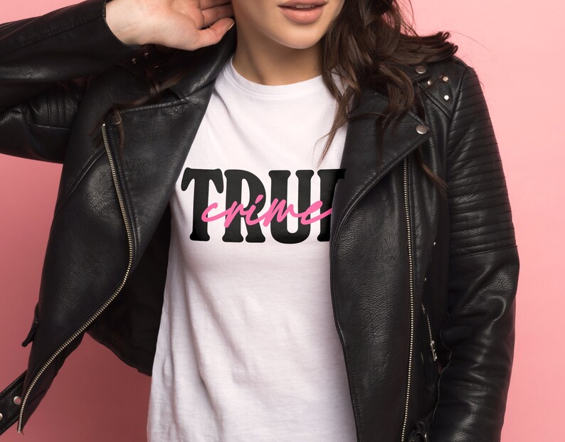 May include: A white t-shirt with black text that says "TRUE crime" in pink lettering. The shirt is being worn by a person wearing a black leather jacket.