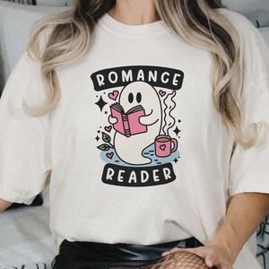 Op de afbeelding: Wit T-shirt met een afbeelding van een geest die een boek leest en een kopje koffie vasthoudt. De tekst "Romance Reader" staat onder de geest.