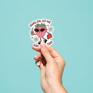 May include: A sticker featuring a cartoon strawberry wearing sunglasses and holding a drink and a purse. The text on the sticker reads "Hang on, let me girl this".