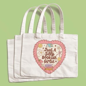 May include: Three white tote bags with long handles. Each bag features a heart-shaped design with the text "Just A Silly Bookish Girlie" and illustrations of a flower, strawberry, and books. The design is in shades of pink, beige, and yellow.