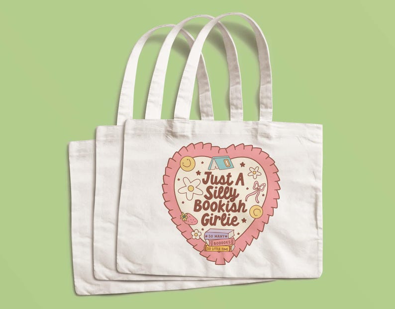 May include: Three white tote bags with long handles. Each bag features a heart-shaped design with the text "Just A Silly Bookish Girlie" and illustrations of a flower, strawberry, and books. The design is in shades of pink, beige, and yellow.