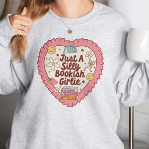 May include: A light gray sweatshirt featuring a heart-shaped graphic with the text "Just a Silly Bookish Girlie." The graphic includes a tent, flowers, a strawberry, a bow, and a stack of books. The sweatshirt is a crew neck style.