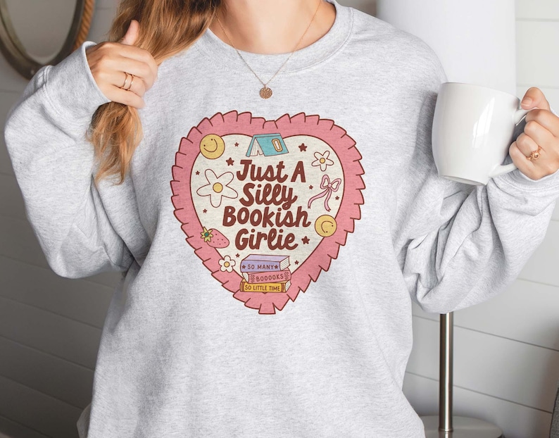 May include: A light gray sweatshirt featuring a heart-shaped graphic with the text "Just a Silly Bookish Girlie." The graphic includes a tent, flowers, a strawberry, a bow, and a stack of books. The sweatshirt is a crew neck style.
