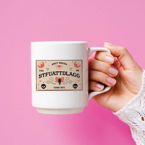 Puede incluir: Taza de cer&aacute;mica blanca con un gr&aacute;fico negro y rojo de una tabla Ouija con el texto "SMUT BOARD" y "STFUATTDLAGG".