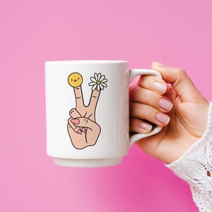 May include: White ceramic mug with a peace sign, a daisy, and a smiley face on the side. The mug is being held by a hand.