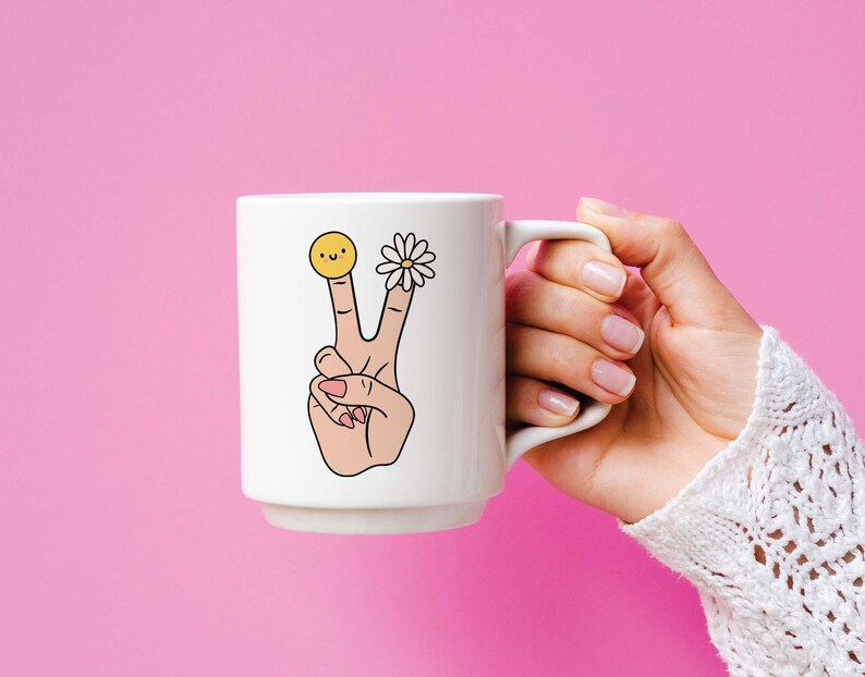 May include: White ceramic mug with a peace sign, a daisy, and a smiley face on the side. The mug is being held by a hand.