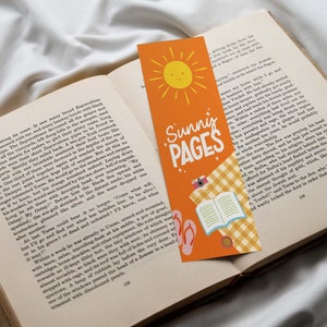May include: Orange bookmark with the text "Sunny Pages" and a smiling sun illustration. The bookmark also features a camera, open book, and flip-flops. The bookmark is placed in an open book.