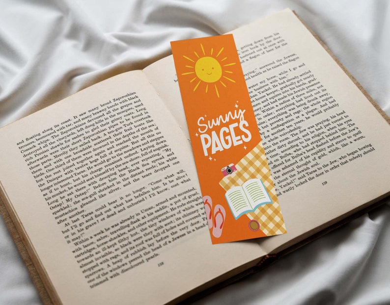 May include: Orange bookmark with the text "Sunny Pages" and a smiling sun illustration. The bookmark also features a camera, open book, and flip-flops. The bookmark is placed in an open book.