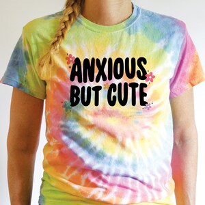 May include: A tie-dye t-shirt with the text "Anxious But Cute" in black lettering. The shirt has a rainbow colour scheme with yellow, orange, pink, blue, and green.