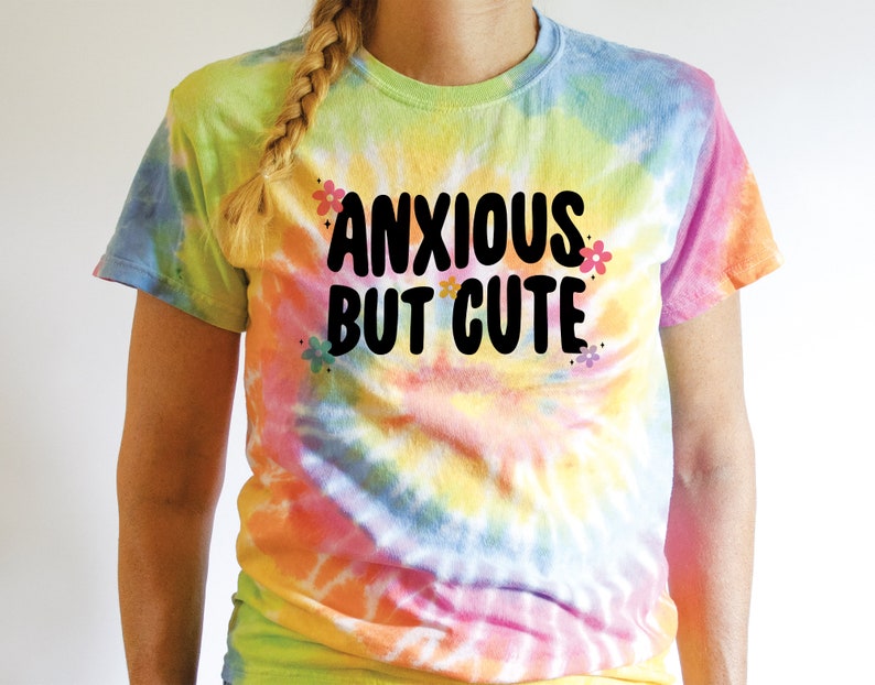 May include: A tie-dye t-shirt with the text "Anxious But Cute" in black lettering. The shirt has a rainbow colour scheme with yellow, orange, pink, blue, and green.
