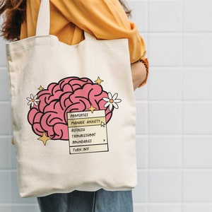 May include: A white canvas tote bag featuring a pink brain illustration and a yellow menu bar with the text "PROPERTIES", "MANAGE ANXIETY", "REFRESH", "TROUBLESHOOT BOUNDARIES", and "TURN OFF".