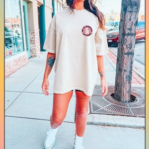 Op de afbeelding: Een beige oversized t-shirt met een roze cirkelvormige afbeelding met de tekst "Not All Disabilities Are Visible". Het model draagt het shirt met een zwarte broek en witte sneakers.