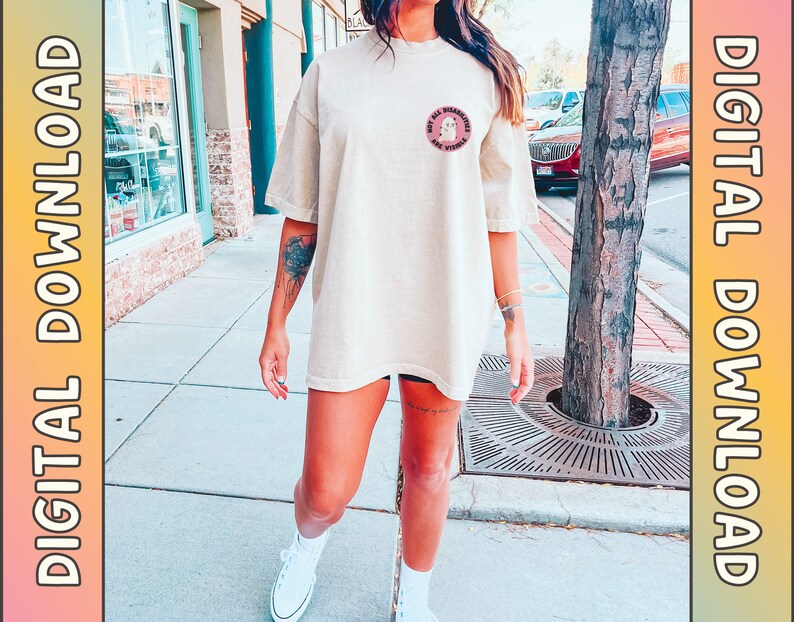 Op de afbeelding: Een beige oversized t-shirt met een roze cirkelvormige afbeelding met de tekst "Not All Disabilities Are Visible". Het model draagt het shirt met een zwarte broek en witte sneakers.
