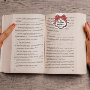 May include: An open book with a bookmark displaying "The Thriller Readers Society" and a red bow. The book is open to pages 406 and 407, with text in Spanish. A hand points to the text on the left page.