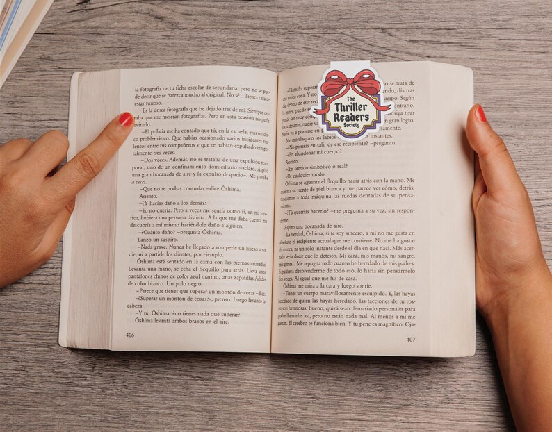 May include: An open book with a bookmark displaying "The Thriller Readers Society" and a red bow. The book is open to pages 406 and 407, with text in Spanish. A hand points to the text on the left page.
