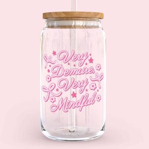 May include: A clear glass water bottle with a pink design that says "Very Demure, Very Mindful". The bottle has a wooden lid and a straw.
