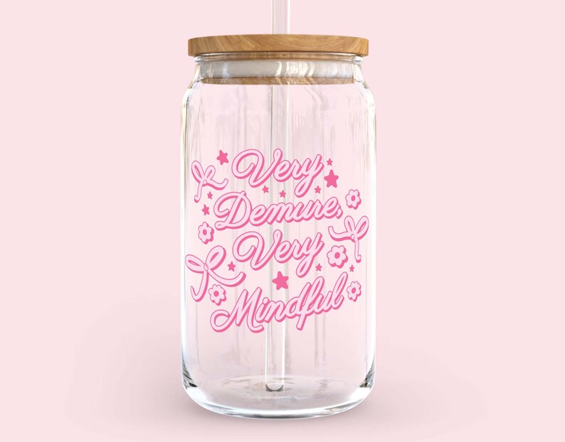 May include: A clear glass water bottle with a pink design that says "Very Demure, Very Mindful". The bottle has a wooden lid and a straw.
