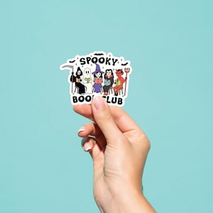Puede incluir: Una pegatina blanca con el texto "Spooky Book Club" e ilustraciones de personajes de Halloween. Los personajes incluyen la Parca, un fantasma, una bruja y un diablo. La pegatina est&aacute; sujeta por una mano sobre un fondo azul claro.
