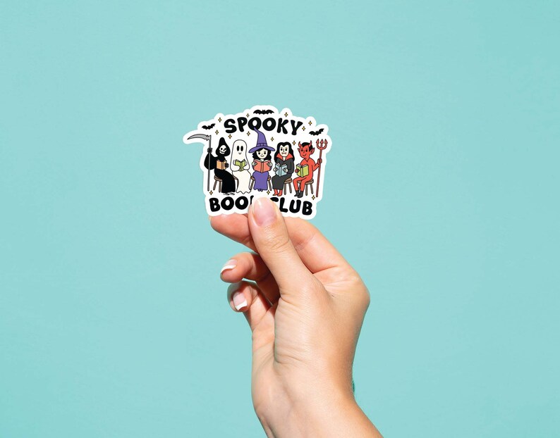 Puede incluir: Una pegatina blanca con el texto "Spooky Book Club" e ilustraciones de personajes de Halloween. Los personajes incluyen la Parca, un fantasma, una bruja y un diablo. La pegatina est&aacute; sujeta por una mano sobre un fondo azul claro.