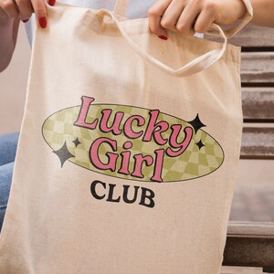 May include: A beige canvas tote bag with a retro-style design featuring the text "Lucky Girl Club" in pink and black lettering inside a green oval with a checkered pattern.