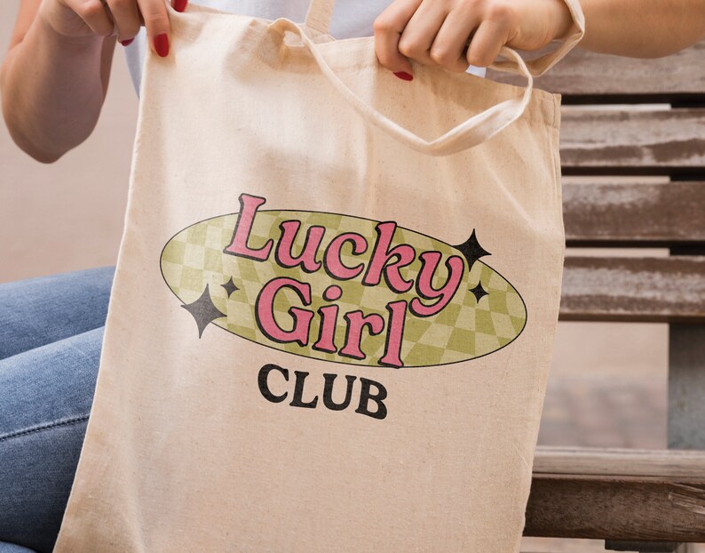 May include: A beige canvas tote bag with a retro-style design featuring the text "Lucky Girl Club" in pink and black lettering inside a green oval with a checkered pattern.