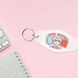 May include: White motel key chain with a pink heart and two books tied together with a ribbon. The text on the keychain reads "Book Besties 4EVER".