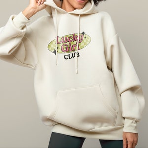 May include: A cream-coloured hoodie with a large graphic print of a retro-style oval with the text "Lucky Girl Club" in pink and green letters.