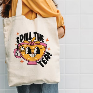 May include: A cream-colored canvas tote bag featuring a graphic illustration of a smiling tea cup with a tea bag inside. The design includes the text "SPILL THE TEA!" in bold black letters, accented by orange star shapes and small white flowers.