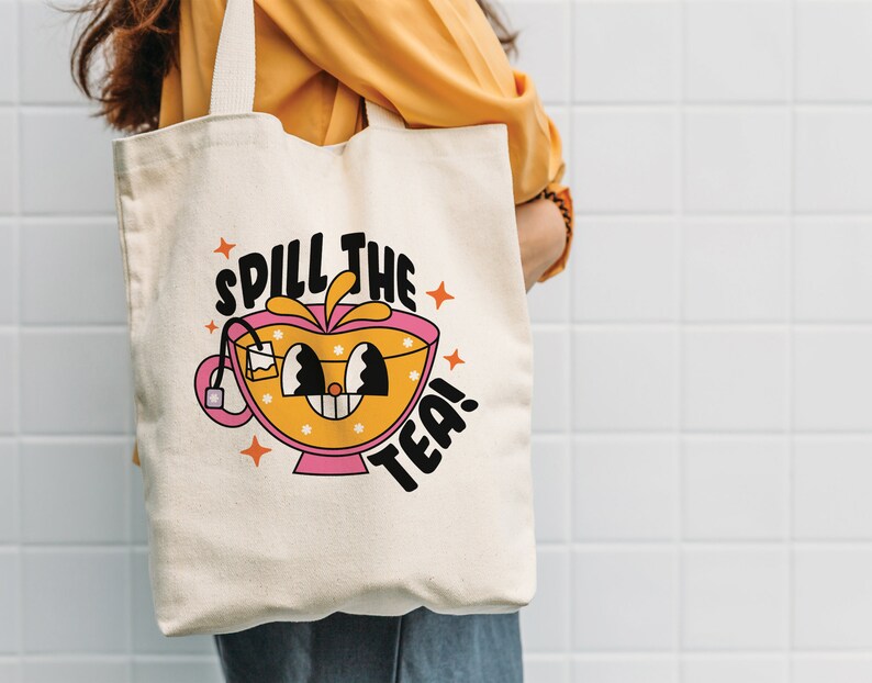 May include: A cream-colored canvas tote bag featuring a graphic illustration of a smiling tea cup with a tea bag inside. The design includes the text "SPILL THE TEA!" in bold black letters, accented by orange star shapes and small white flowers.
