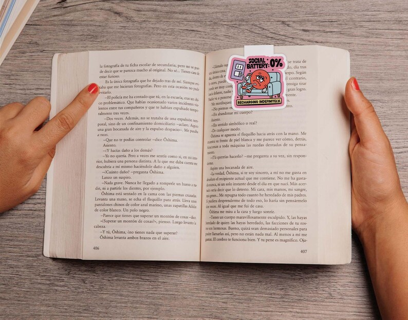 May include: A book is open to page 406-407, with a bookmark in the shape of a sticker. The bookmark features a cartoon of a couch with a person on it, and the text "SOCIAL BATTERY: 0% RECHARGING INDEFINITELY".