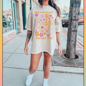 Puede incluir: Una camiseta beige de gran tama&ntilde;o con un estampado gr&aacute;fico de flores de colores, estrellas y un signo de paz. El texto de la camiseta dice "Have a great day! You got this!"