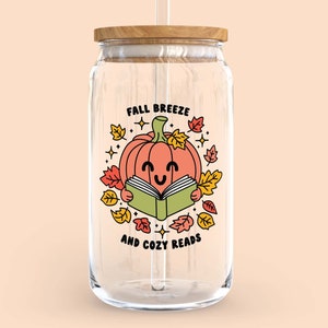 May include: A clear glass jar with a wooden lid and a straw. The jar has a graphic of a smiling pumpkin reading a book with the text "Fall Breeze and Cozy Reads".