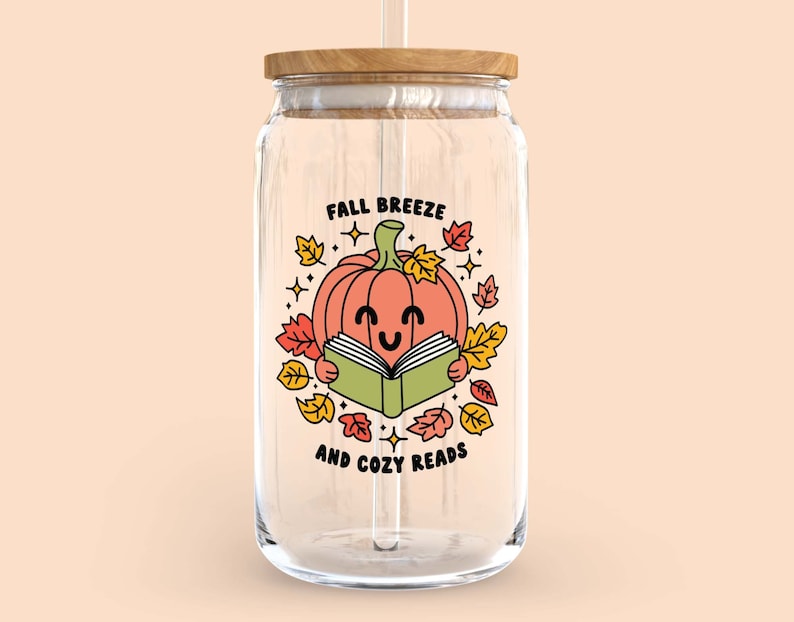May include: A clear glass jar with a wooden lid and a straw. The jar has a graphic of a smiling pumpkin reading a book with the text "Fall Breeze and Cozy Reads".