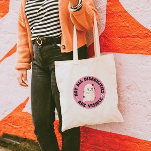 Op de afbeelding: Een witte tote bag met een roze cirkelvormig ontwerp met een spook en de tekst "Not all disabilities are visible".