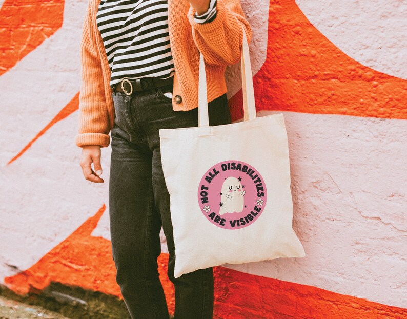Op de afbeelding: Een witte tote bag met een roze cirkelvormig ontwerp met een spook en de tekst "Not all disabilities are visible".
