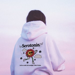 Może przedstawiać: Biała bluza z kapturem z grafiką uśmiechniętego słońca w okularach przeciwsłonecznych, kt&oacute;re biegnie, i tekstem "Serotonin." Grafika jest otoczona motylami, chmurami i kwiatami. Tekst pod grafiką brzmi "Go for a walk. Get sunlight. Take it slow."
