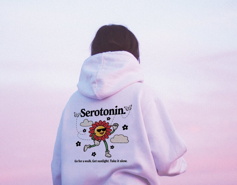 Może przedstawiać: Biała bluza z kapturem z grafiką uśmiechniętego słońca w okularach przeciwsłonecznych, kt&oacute;re biegnie, i tekstem "Serotonin." Grafika jest otoczona motylami, chmurami i kwiatami. Tekst pod grafiką brzmi "Go for a walk. Get sunlight. Take it slow."