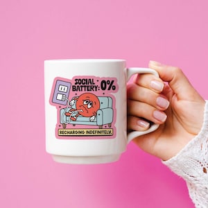 May include: White ceramic mug with a cartoon graphic of a red character on a blue couch with the text "SOCIAL BATTERY: 0% RECHARGING INDEFINITELY". The mug is held against a pink background.