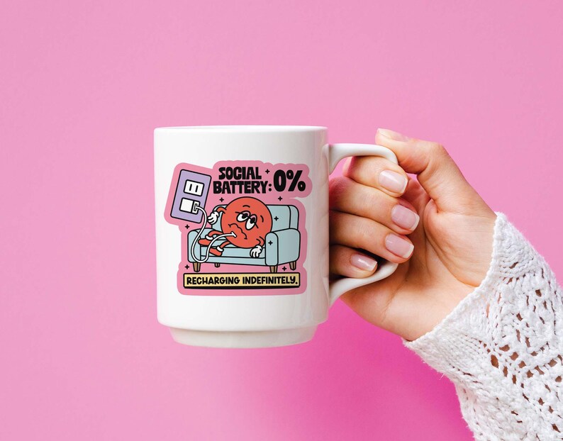 May include: White ceramic mug with a cartoon graphic of a red character on a blue couch with the text "SOCIAL BATTERY: 0% RECHARGING INDEFINITELY". The mug is held against a pink background.