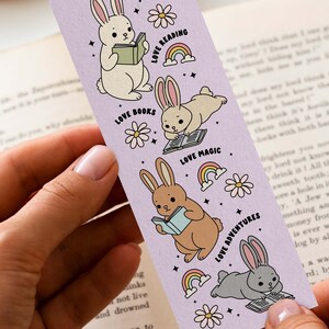 May include: A purple bookmark with illustrations of bunnies reading books. The bookmark has the text "Love Reading", "Love Books", "Love Magic", and "Love Adventures".