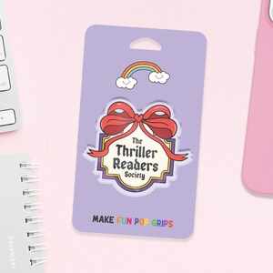 May include: A purple pop grip featuring a red bow and the words "The Thriller Readers Society." A rainbow and clouds graphic is above the bow. The text "MAKE FUN POP GRIPS" is at the bottom.