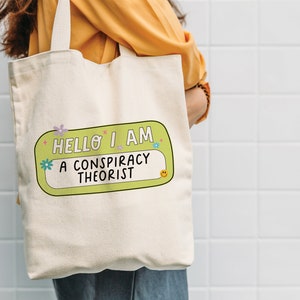 May include: A white canvas tote bag with a green and yellow name tag that says "Hello I am a conspiracy theorist".