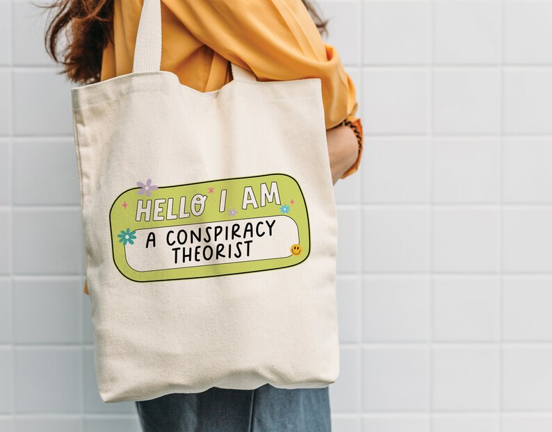May include: A white canvas tote bag with a green and yellow name tag that says "Hello I am a conspiracy theorist".