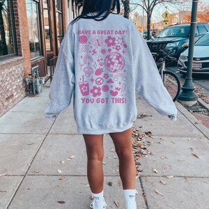 Puede incluir: Sudadera gris con un estampado gr&aacute;fico rosa en la espalda. El gr&aacute;fico incluye el texto "Have a Great Day! You Got This!" y varias ilustraciones rosas de flores, estrellas, corazones y otros dise&ntilde;os.