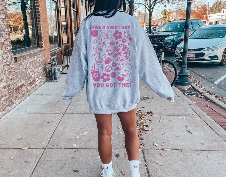Puede incluir: Sudadera gris con un estampado gr&aacute;fico rosa en la espalda. El gr&aacute;fico incluye el texto "Have a Great Day! You Got This!" y varias ilustraciones rosas de flores, estrellas, corazones y otros dise&ntilde;os.