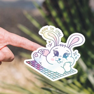 May include: A white bunny with pink ears and a rainbow gradient is lying on a checkered blanket. The bunny has a happy expression and is surrounded by sparkles. The text "Born to Chill" is written in a rainbow gradient above the bunny.