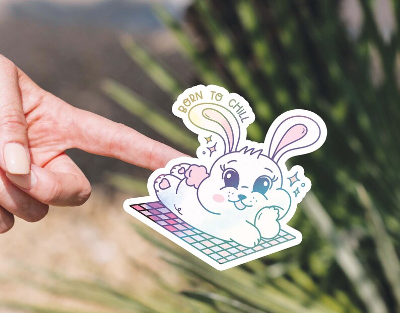 May include: A white bunny with pink ears and a rainbow gradient is lying on a checkered blanket. The bunny has a happy expression and is surrounded by sparkles. The text "Born to Chill" is written in a rainbow gradient above the bunny.