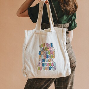 May include: A white tote bag with a colorful graphic that reads "Be the reason someone believes in the goodness of people".
