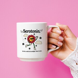 Może przedstawiać: Biały kubek ceramiczny z czarno-białym rysunkiem uśmiechniętego kwiatu w okularach przeciwsłonecznych i napisem "Serotonina" czarnym kolorem. Kubek ma napis "Go for a walk. Get sunlight. Take it slow." nadrukowany na spodzie.